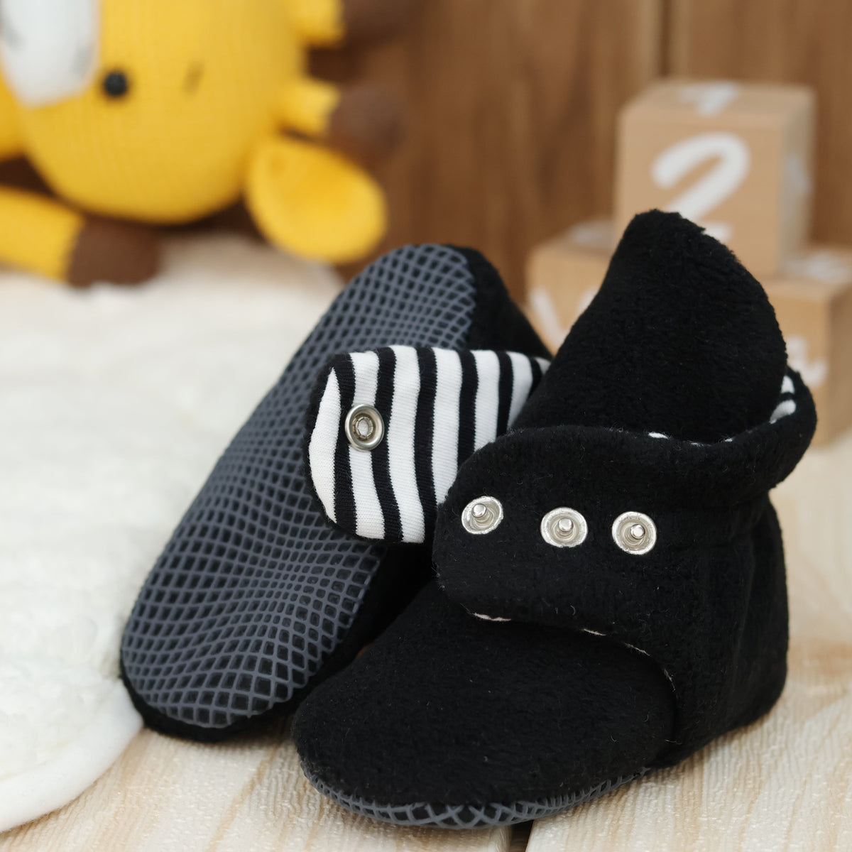 Close-up of black fleece baby booties with striped lining and snap straps, showing one sole and an open snap strap.