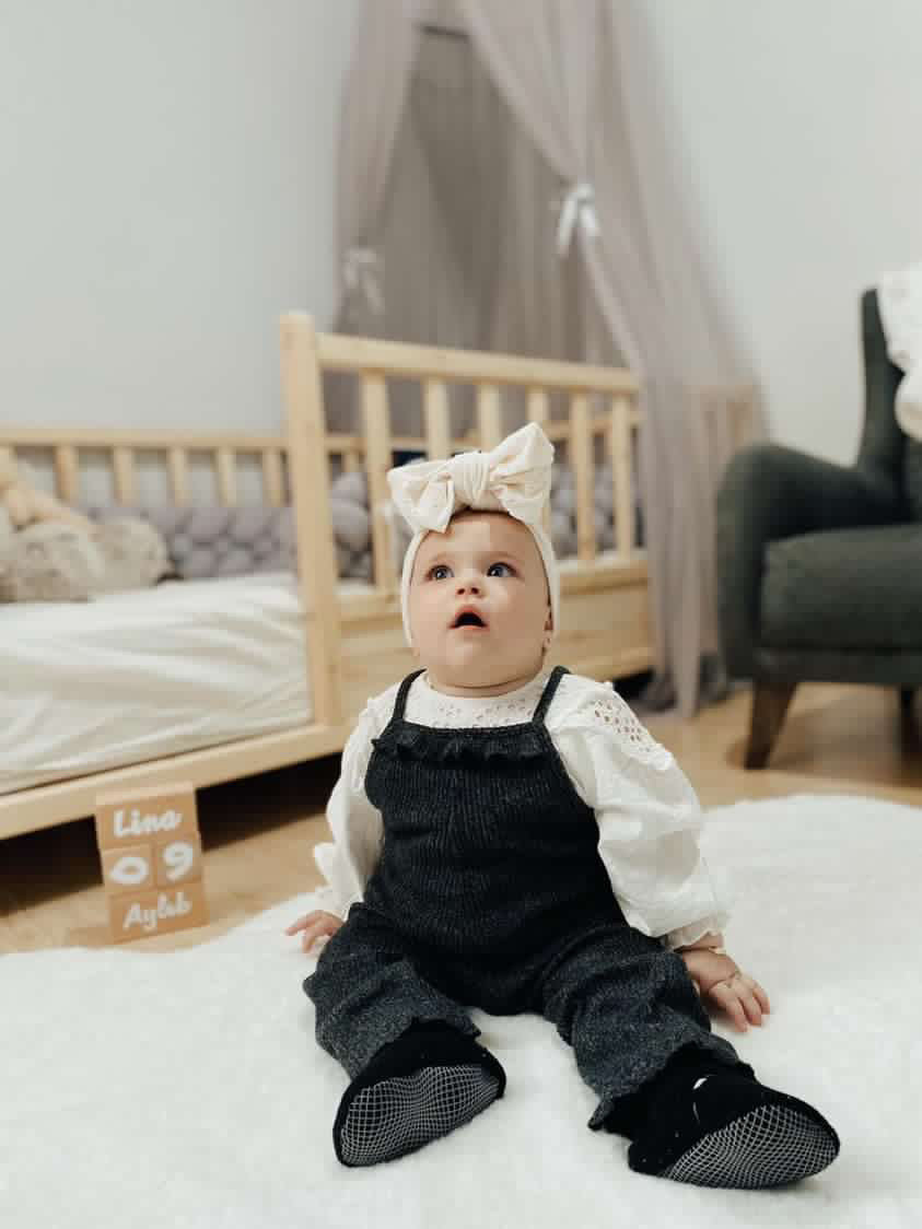 Baby seated on a rug wearing black overalls and black booties.