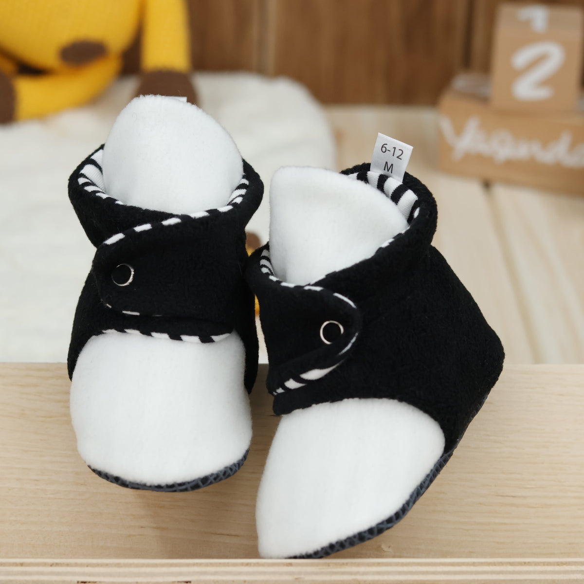 Front view of a pair of black-and-white fleece baby booties with striped lining and snap straps.