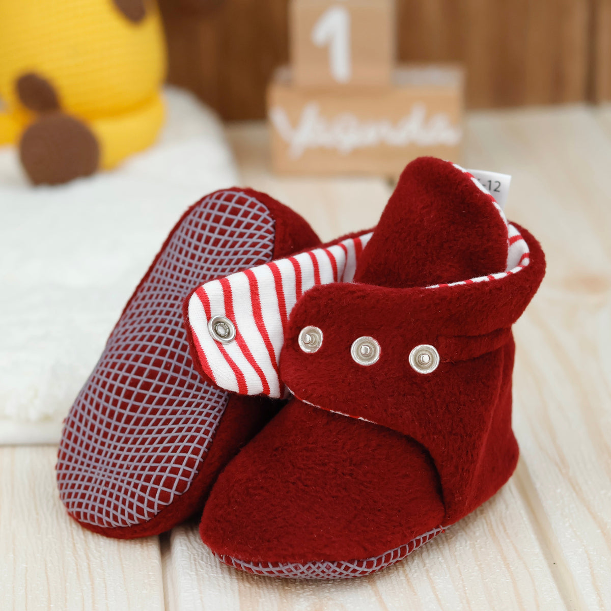 Close-up of burgundy fleece baby booties with striped lining and snap straps, showing one sole and an open snap strap.