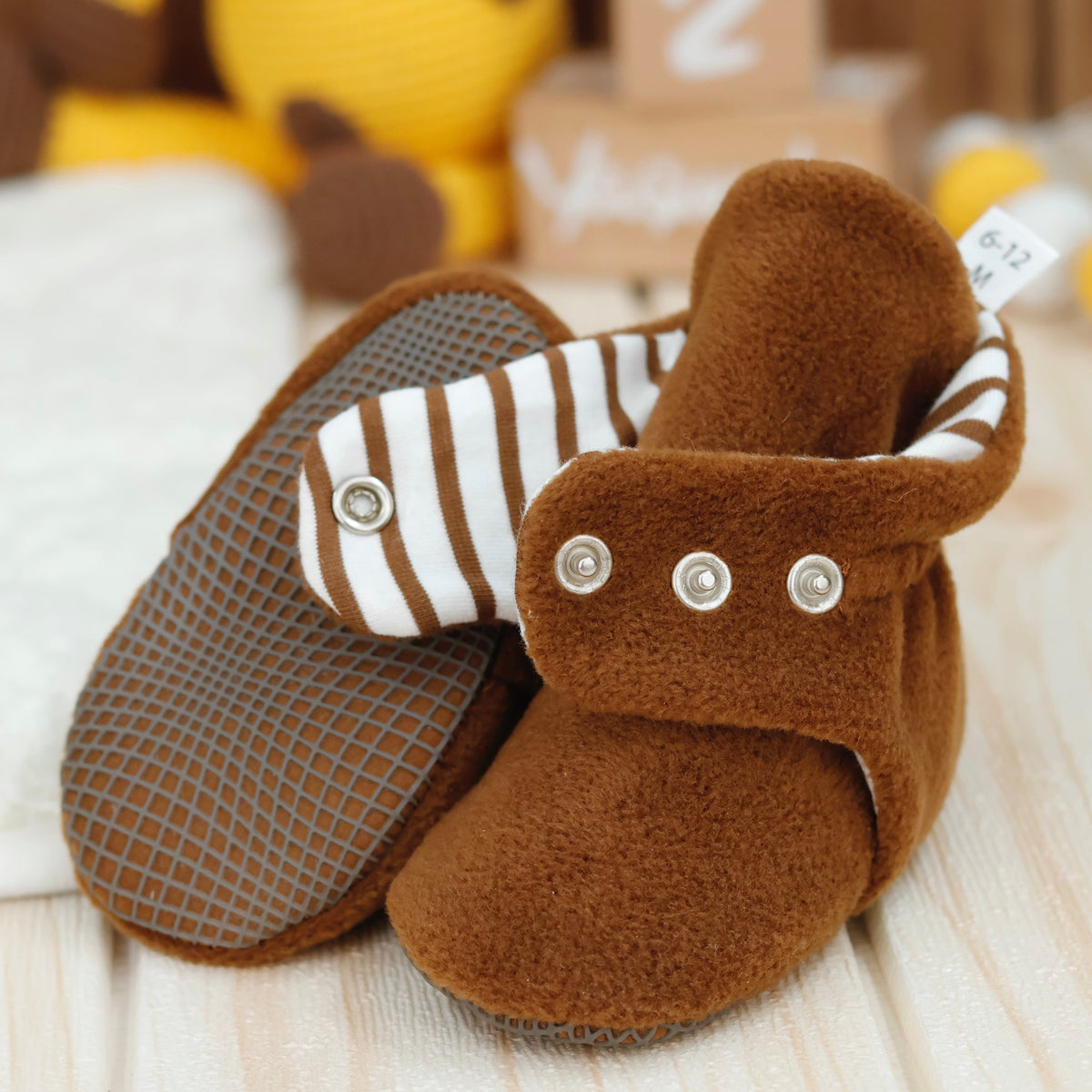 Close-up of warm brown fleece baby booties with striped lining and snap straps, showing one sole and an open snap strap.