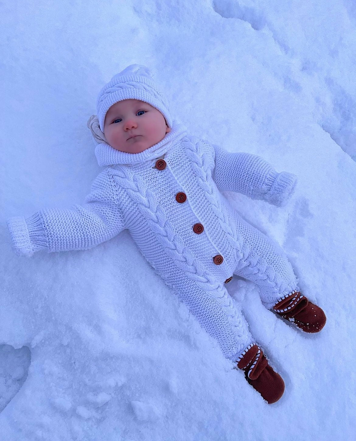 Baby in a white outfit lying in snow wearing brown booties.