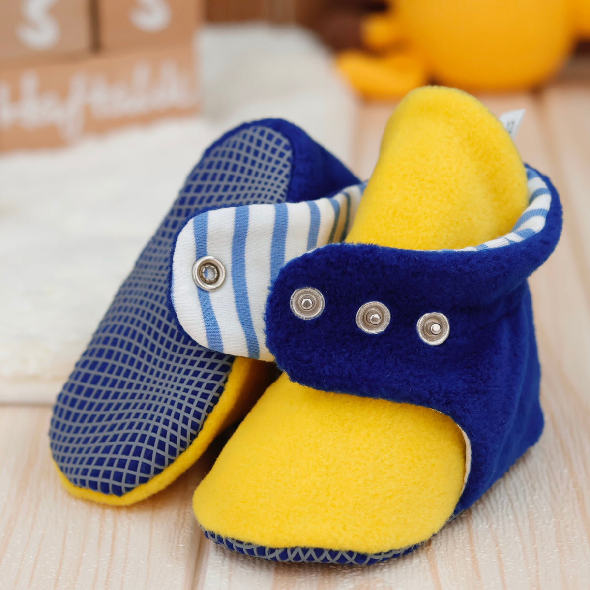Close-up of blue-and-yellow fleece baby booties with striped lining and snap straps, showing one sole and an open snap strap.
