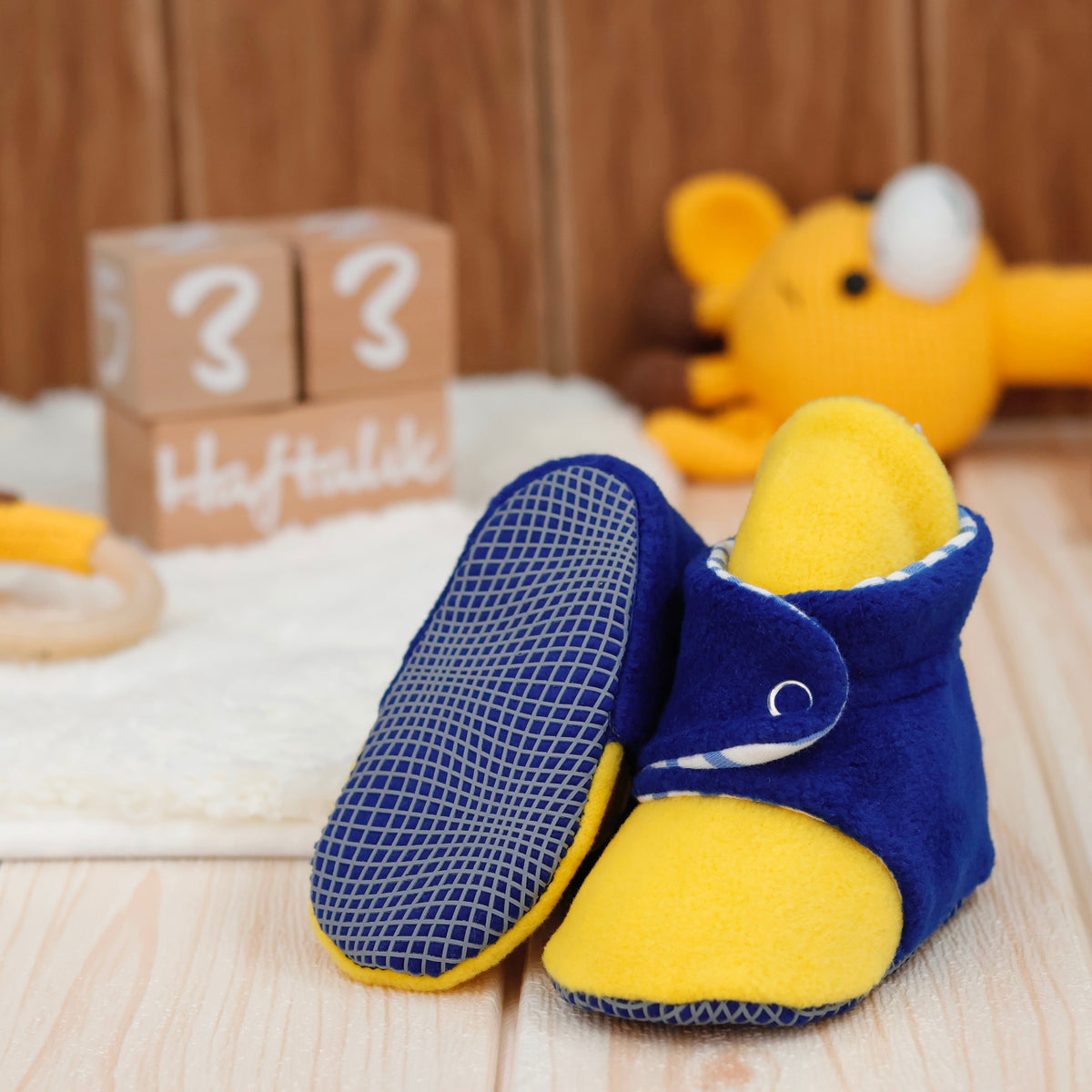Blue-and-yellow fleece baby booties with striped lining and snap straps arranged on a surface with one sole facing the camera.