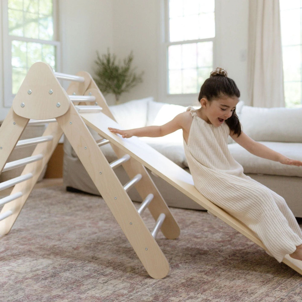 Child sitting near the lower end of a smooth ramp attached to a white-rung climbing triangle.