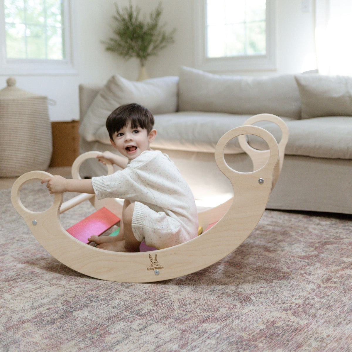 Child seated in a rainbow-slat rocker, holding a circular handle in a living room.
