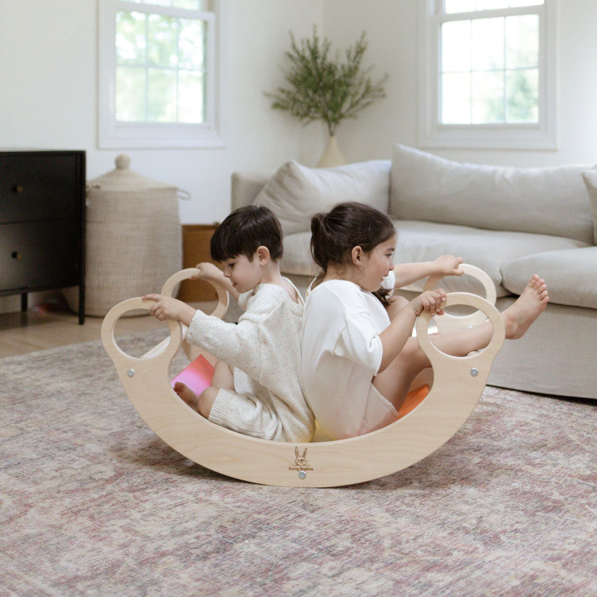 Two children seated back to back in a rainbow-slat rocker in a living room.
