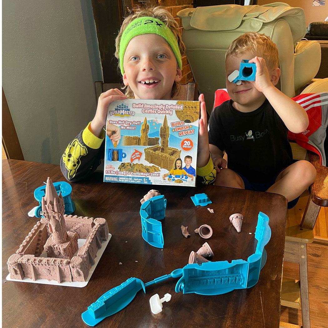 Two children at a table with a BuildMaster kit box, blue molds, roof pieces and a molded sand castle.