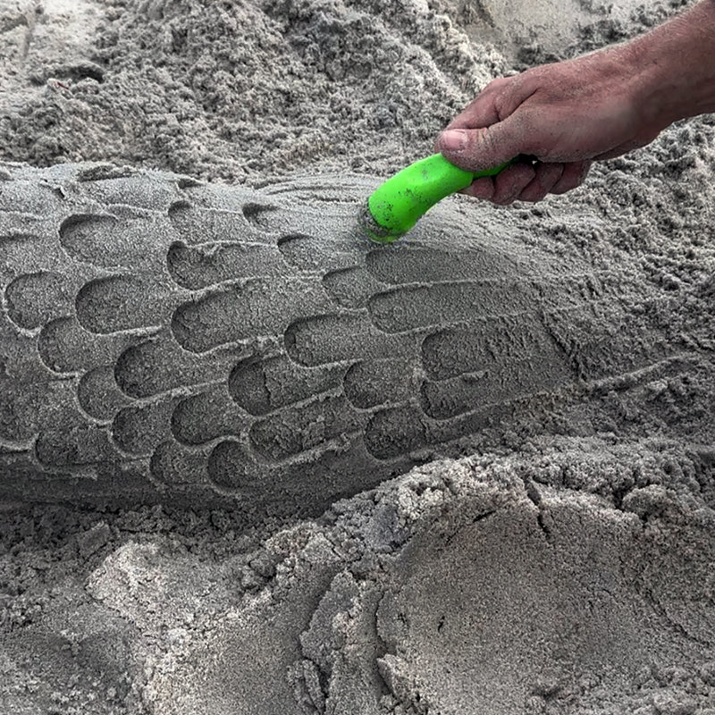 Hand using a green sculpting tool to carve overlapping scale shapes into a sand mound.