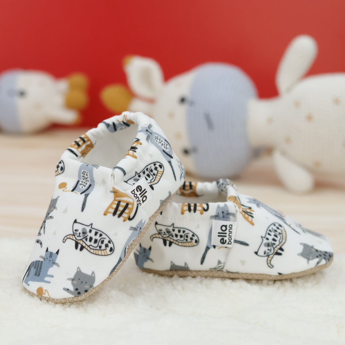 Cream slip-on baby moccasins with illustrated cat print arranged together to show the sides and uppers.