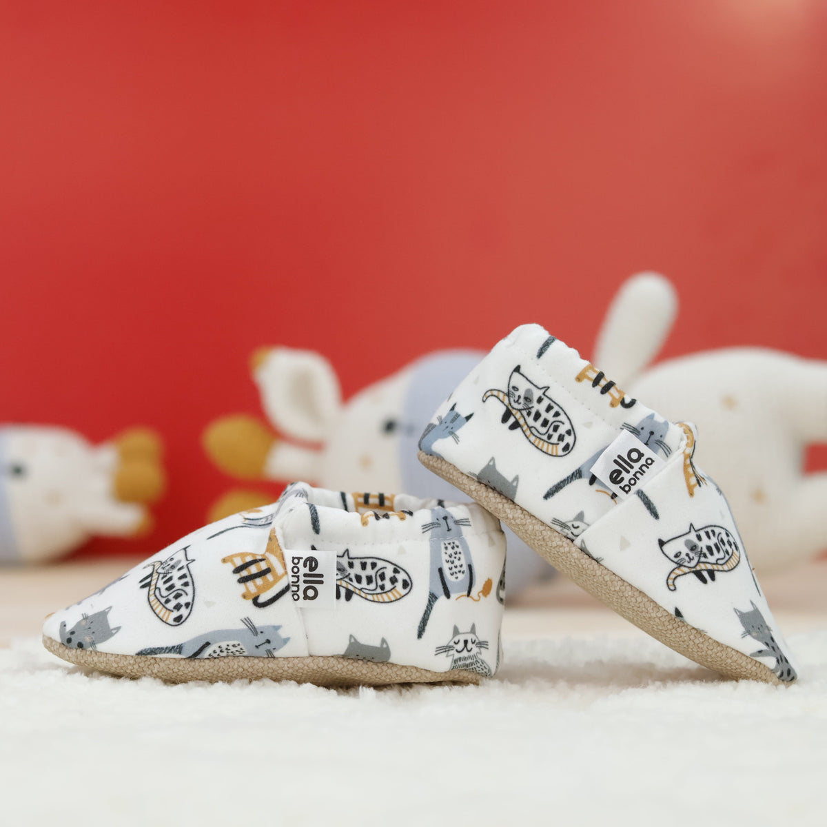 Cream slip-on baby moccasins with illustrated cat print arranged together to show the sides and uppers.