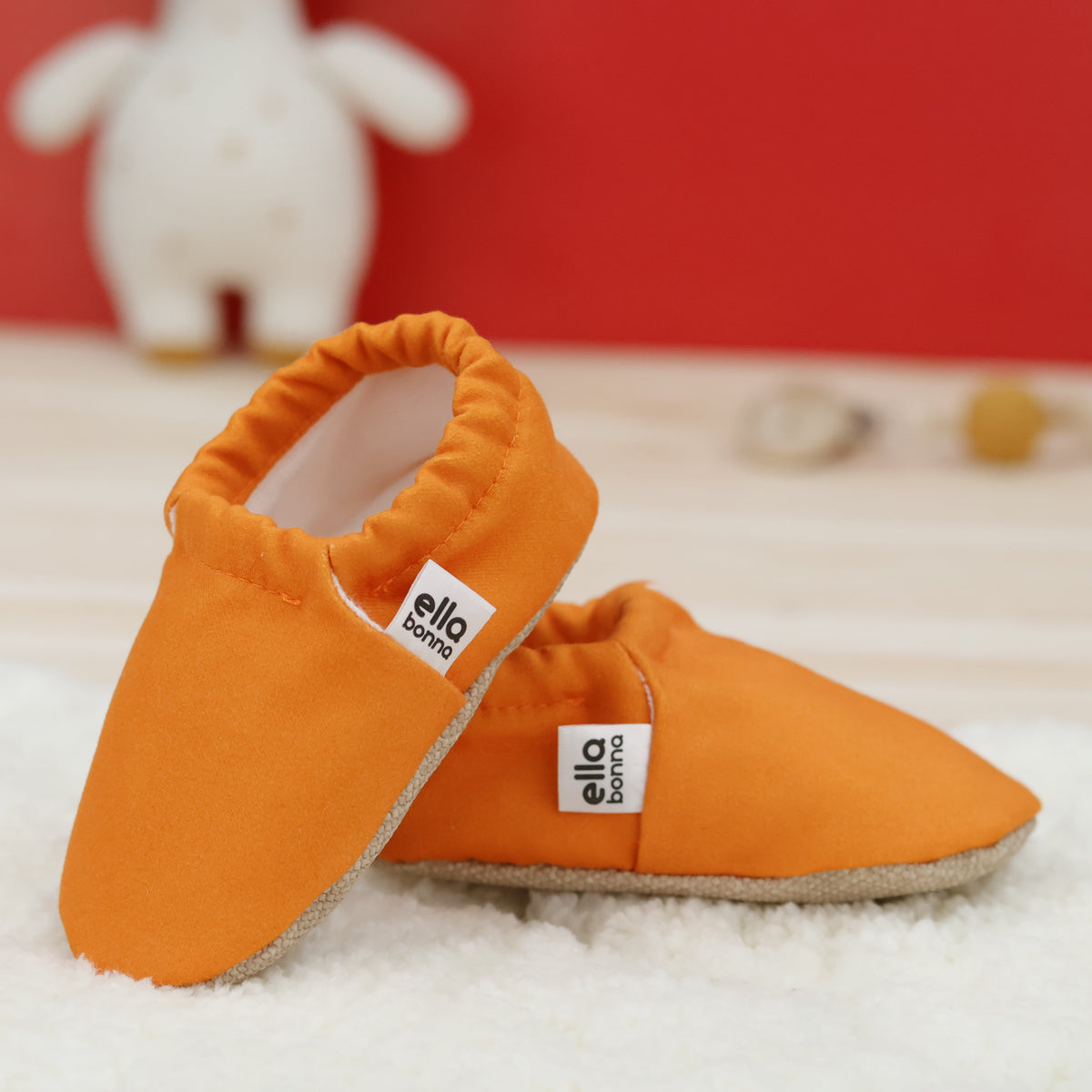 Orange slip-on baby moccasins with cream lining and tan soles arranged together to show the sides and uppers.