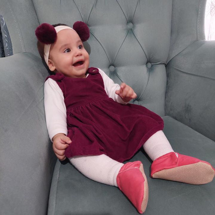 Baby seated in a gray chair wearing pink moccasins.