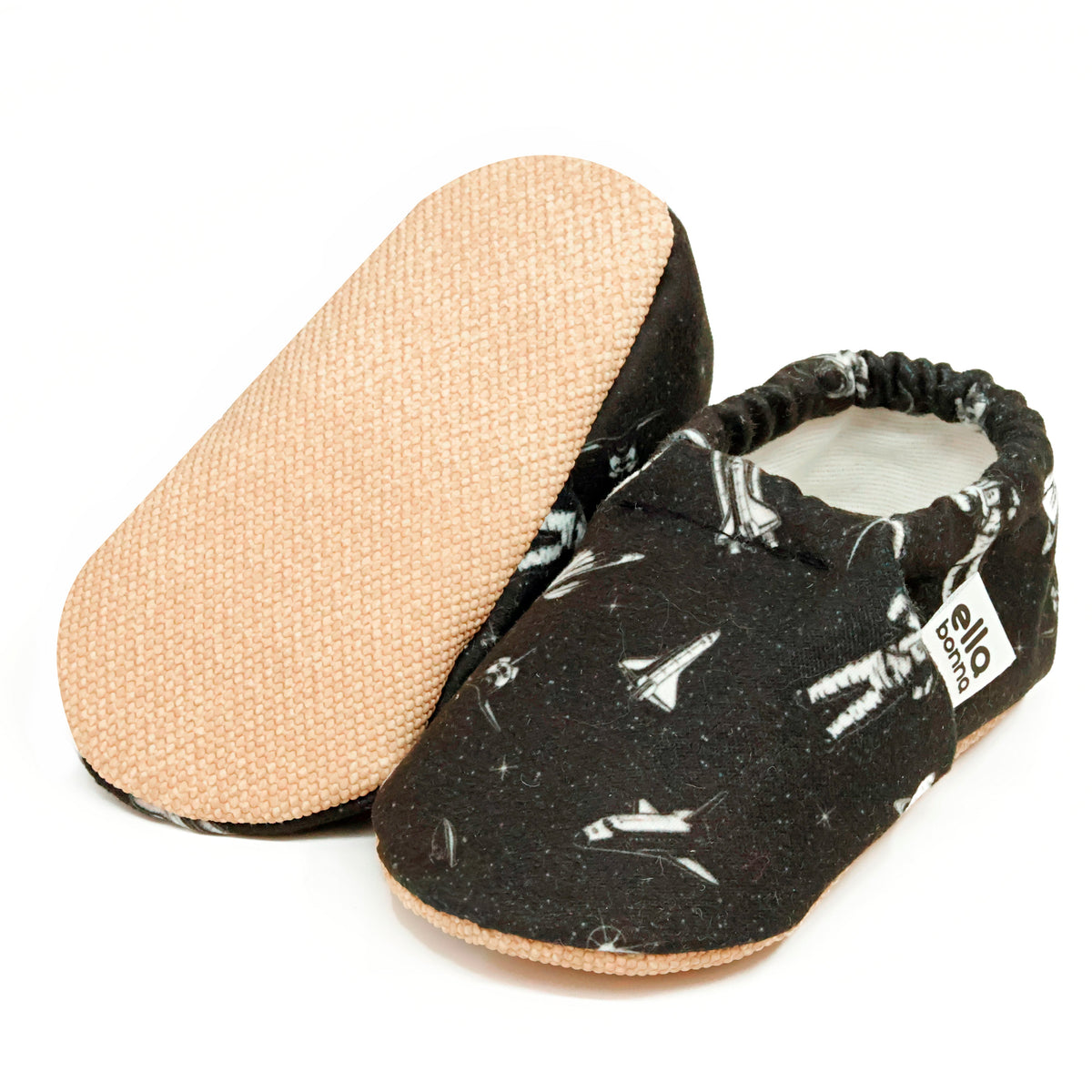 Black slip-on baby moccasins with white astronaut and spacecraft print, with one sole facing the camera on a white background.