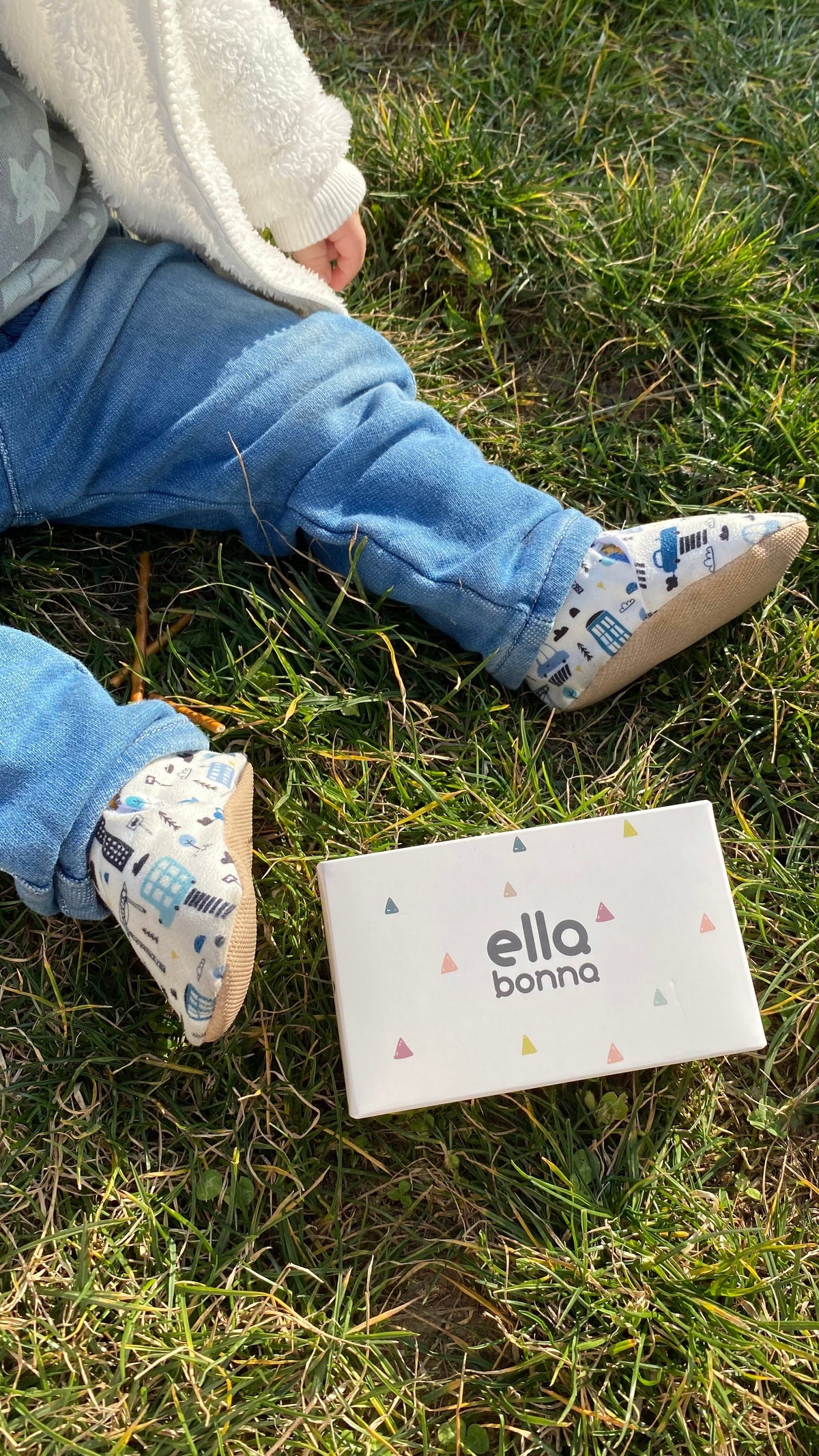 Child seated on grass with city-print moccasins and an Ella Bonna card.