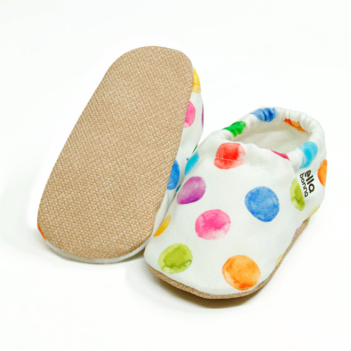 White slip-on baby moccasins with multicolored watercolor dot print, with one sole facing the camera on a white background.