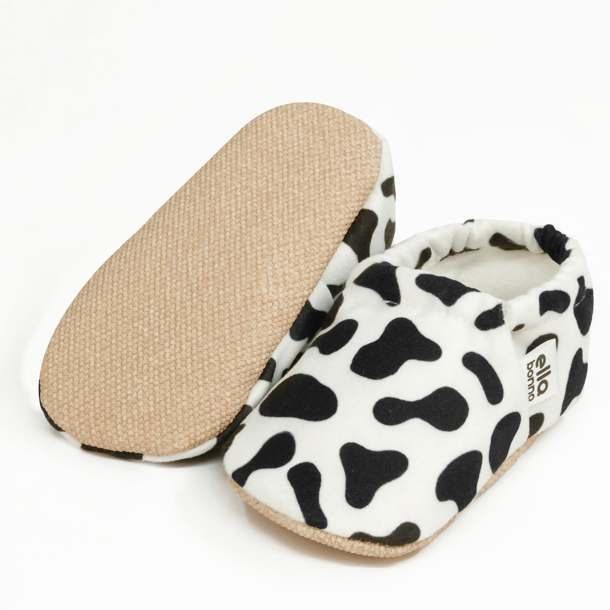 White slip-on baby moccasins with black cow spots, with one sole facing the camera on a white background.