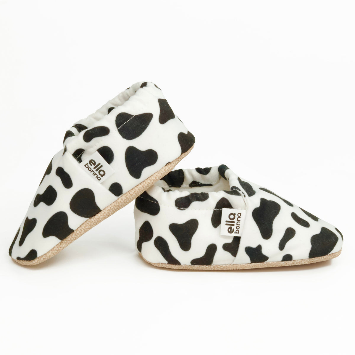 Side views of white slip-on baby moccasins with black cow spots on a white background.