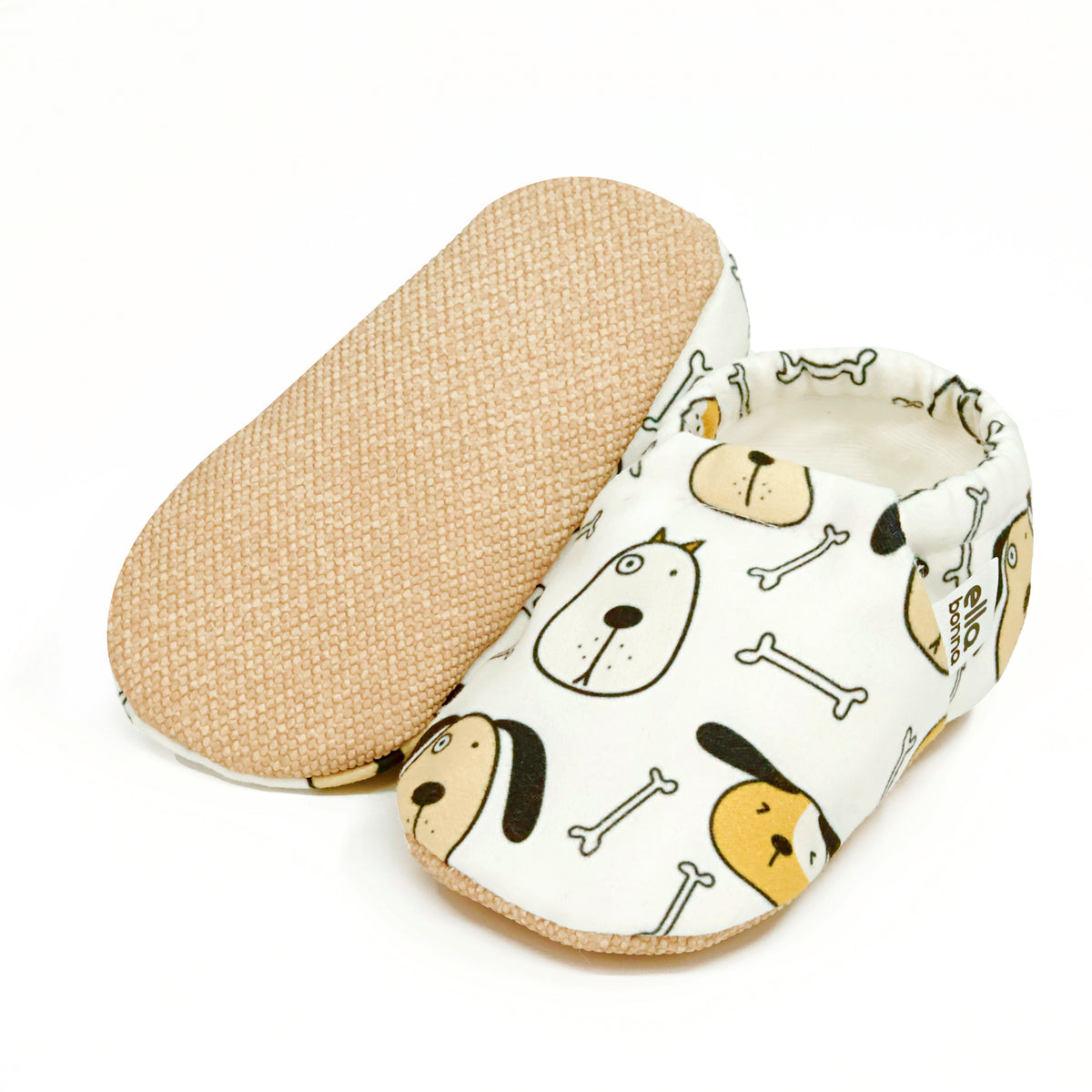 Cream slip-on baby moccasins with dog faces and bone print, with one sole facing the camera on a white background.