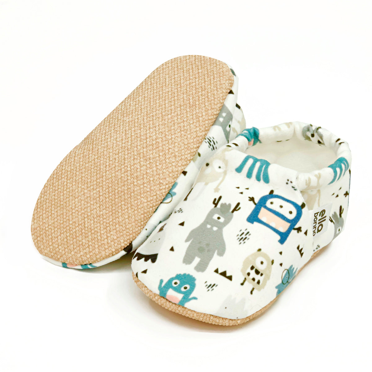 Cream slip-on baby moccasins with blue-and-gray cartoon monster print, with one sole facing the camera on a white background.