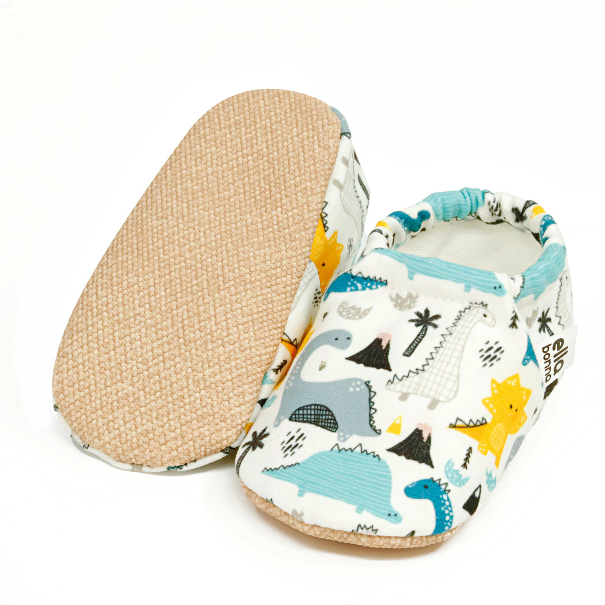 Cream slip-on baby moccasins with blue, gray and yellow dinosaur print, with one sole facing the camera on a white background.