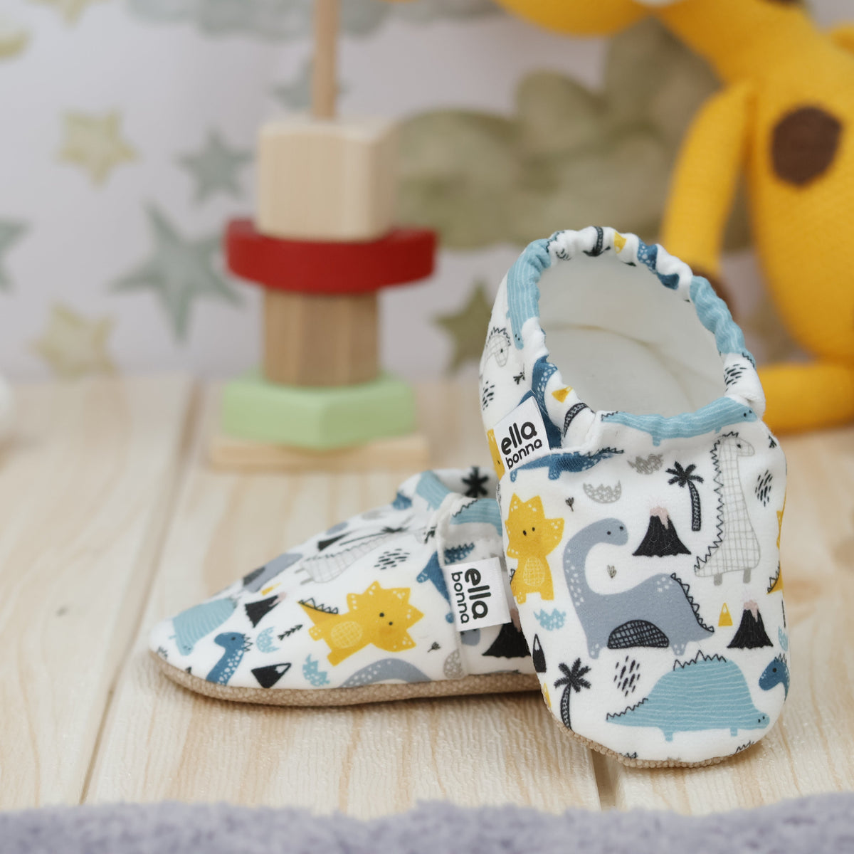 Cream slip-on baby moccasins with blue, gray and yellow dinosaur print arranged together to show the sides and uppers.