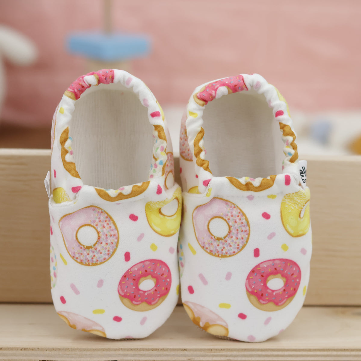 Pair of cream slip-on baby moccasins with pink-and-yellow doughnut print, showing the openings and uppers.