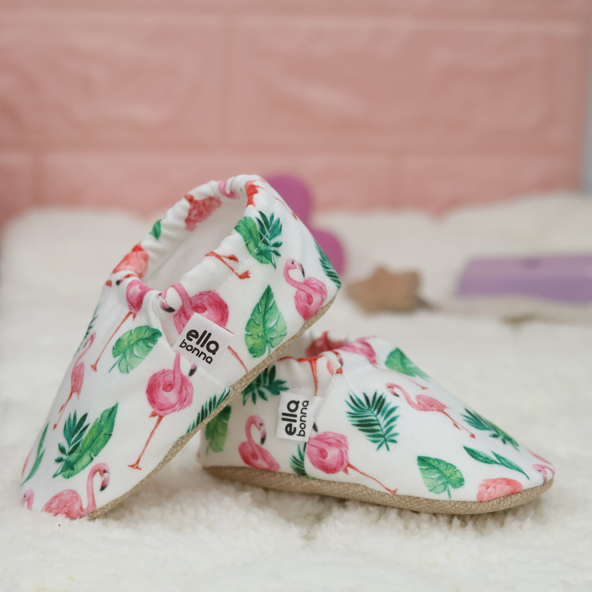 Cream slip-on baby moccasins with pink flamingos and green leaves arranged together to show the sides and uppers.