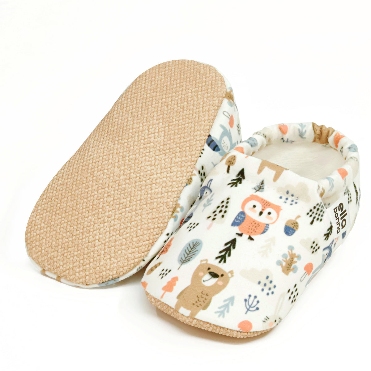 Cream slip-on baby moccasins with woodland animal and tree print, with one sole facing the camera on a white background.
