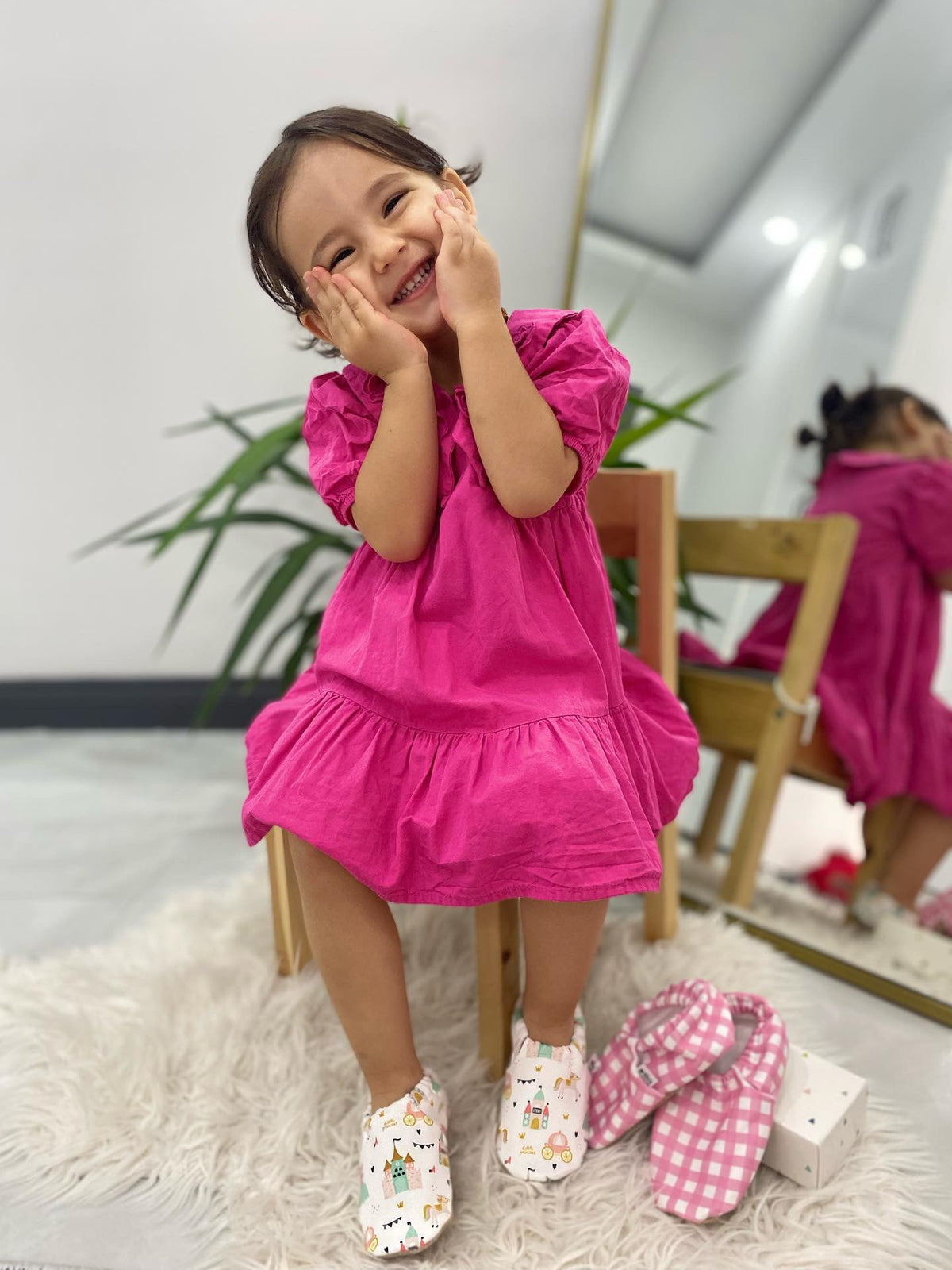 Child in a pink dress seated with princess-print and gingham moccasins visible.