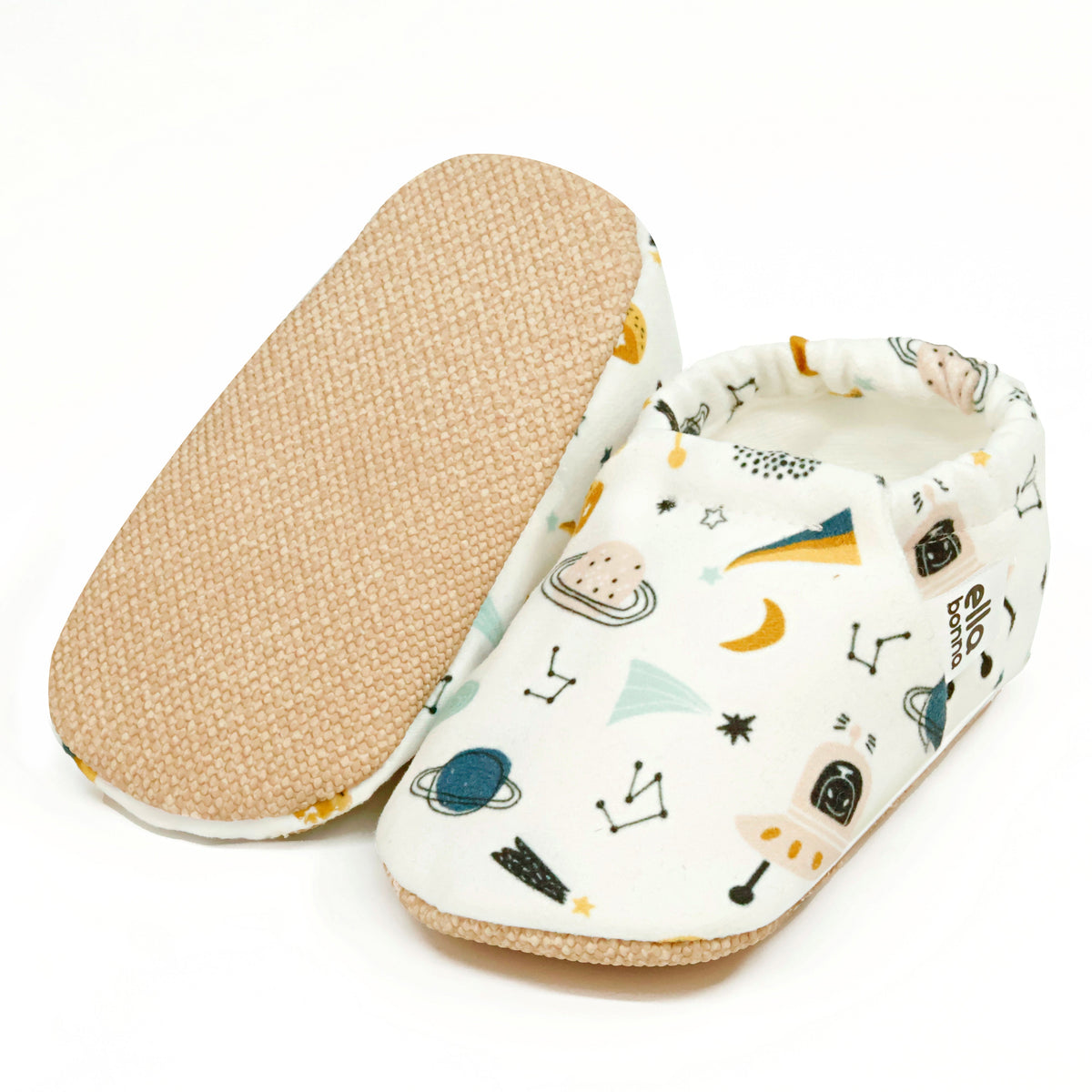 Cream slip-on baby moccasins with planets, rockets and constellation print, with one sole facing the camera on a white background.