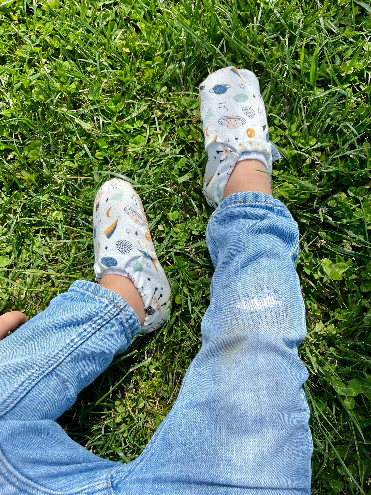 Overhead view of space-print moccasins worn with jeans on grass.
