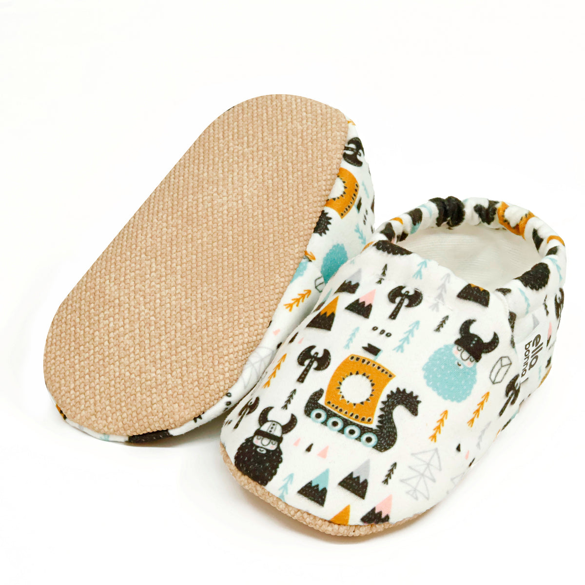 Cream slip-on baby moccasins with Viking ship and mountain print, with one sole facing the camera on a white background.