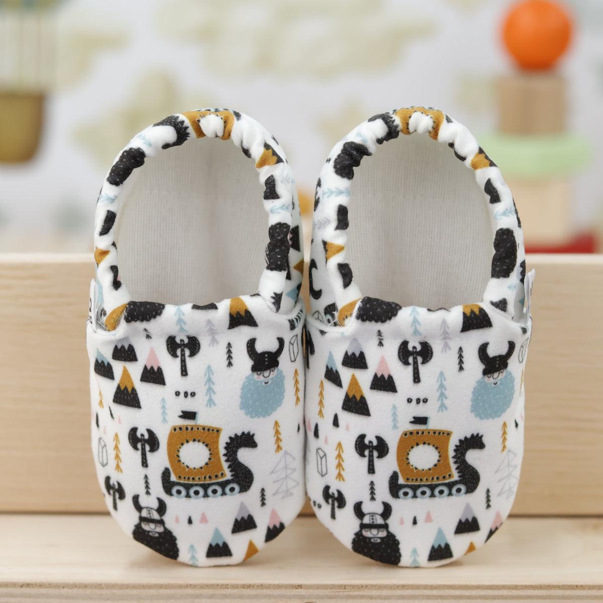 Pair of cream slip-on baby moccasins with Viking ship and mountain print, showing the openings and uppers.