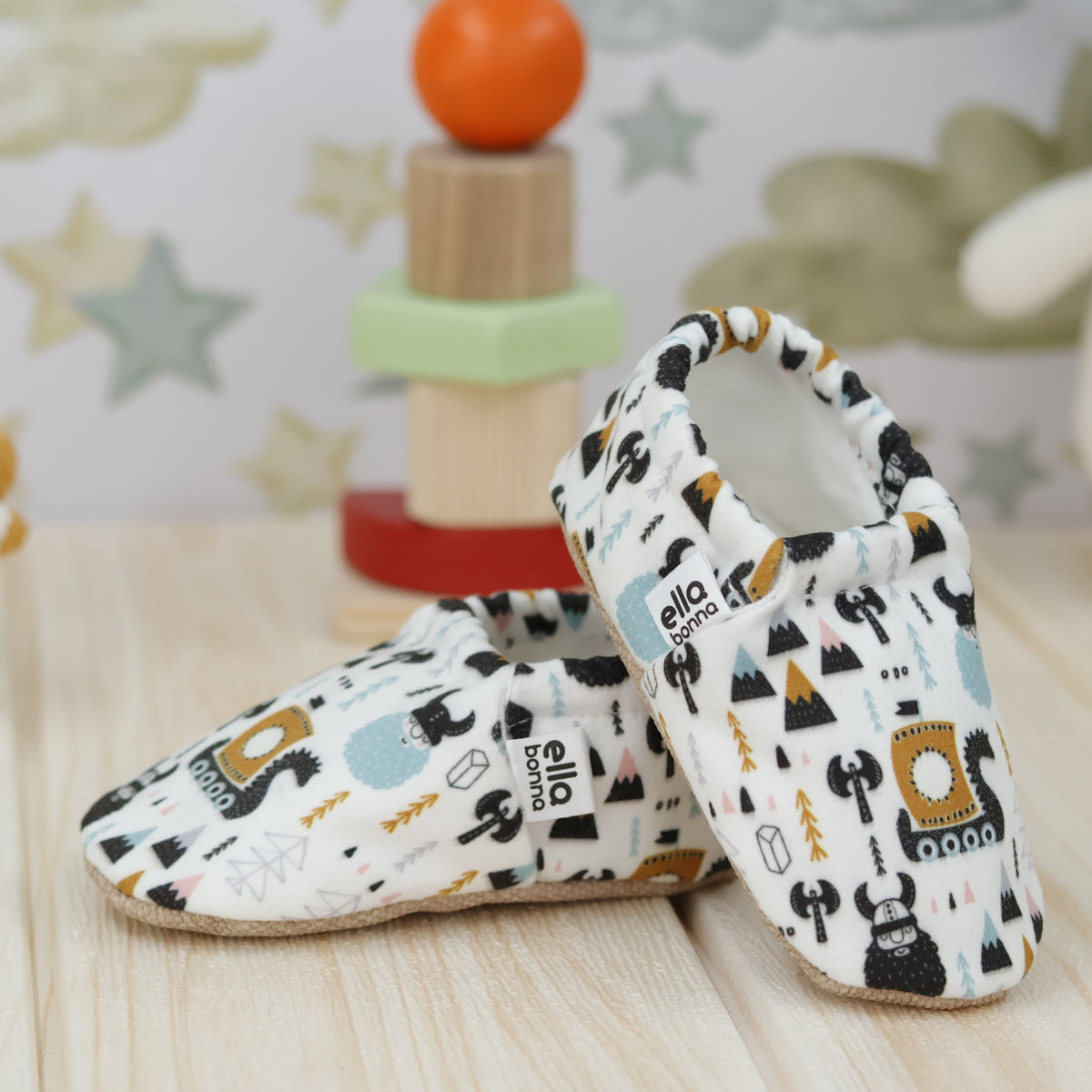 Cream slip-on baby moccasins with Viking ship and mountain print arranged together to show the sides and uppers.