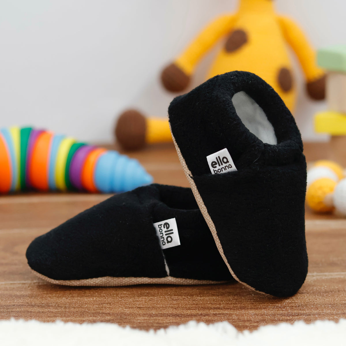 Black fleece slip-on baby moccasins with tan soles arranged together to show the sides and uppers.