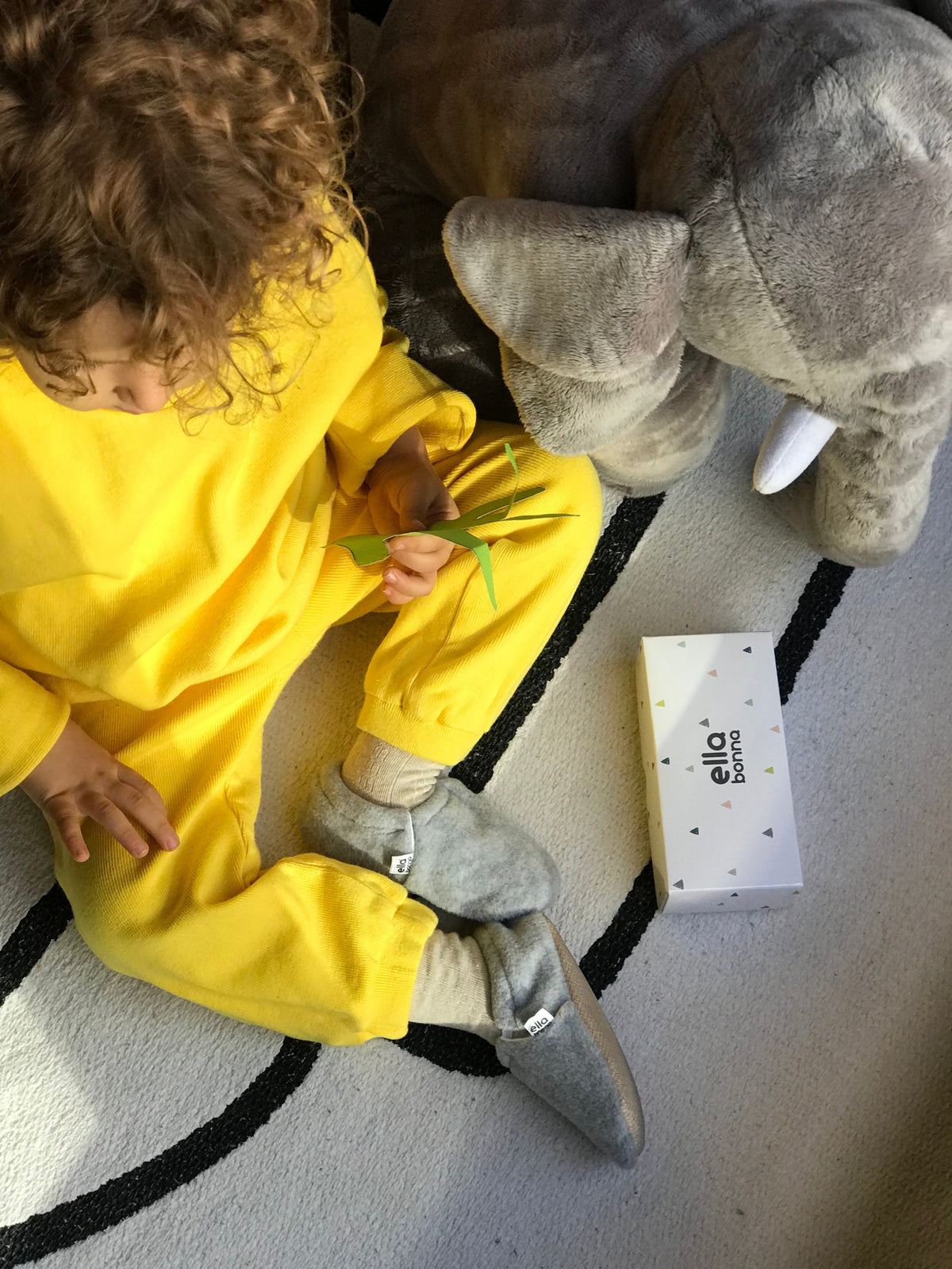 Overhead view of a child wearing gray fleece moccasins beside an Ella Bonna card.