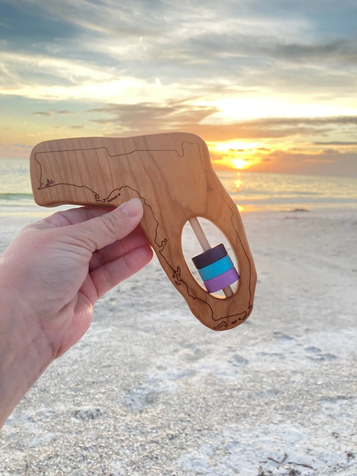 Hand holding a Florida-shaped wooden rattle in front of a beach sunset