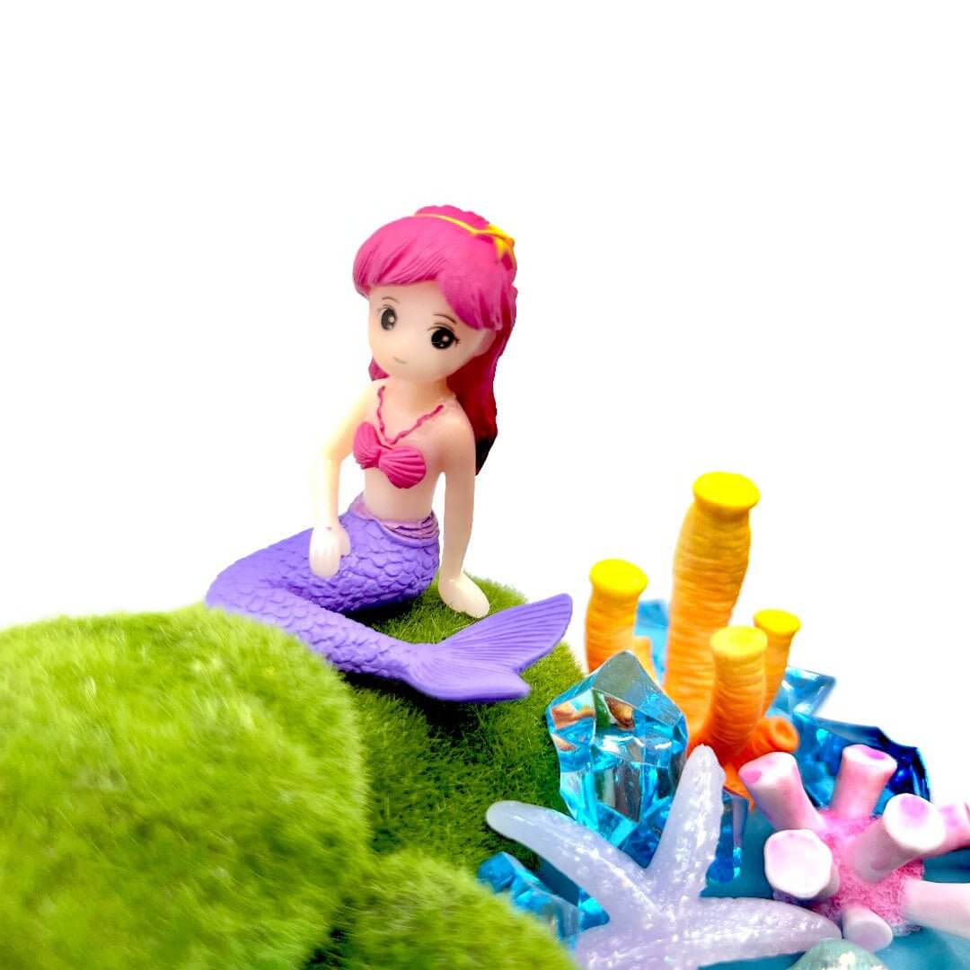 Pink-haired mermaid figure on a green decoration beside coral shapes and blue gems