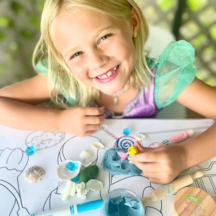Child playing with blue playdough, shells and miniature accessories on an illustrated mat