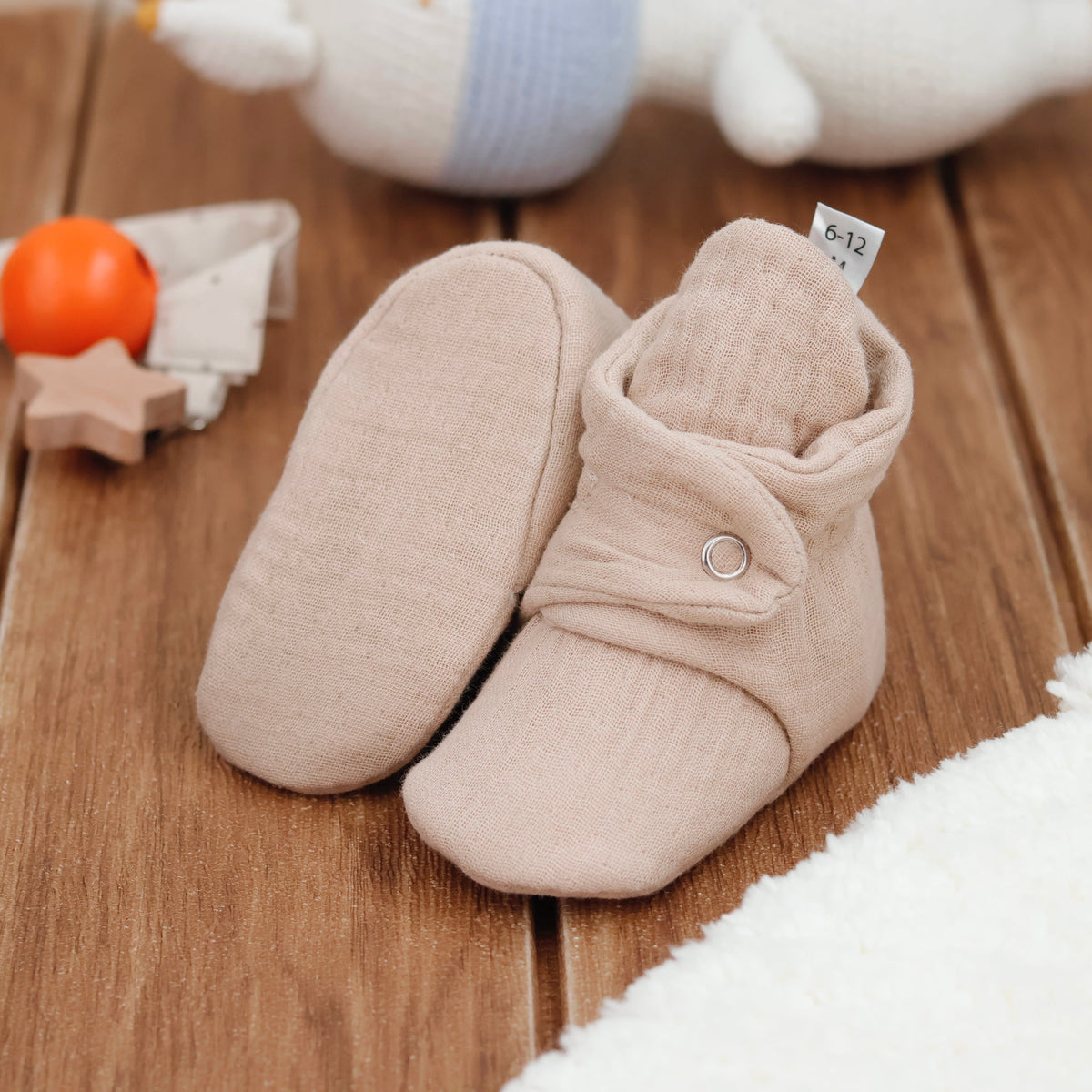 Beige textured muslin baby booties with snap straps arranged on a surface with one sole facing the camera.
