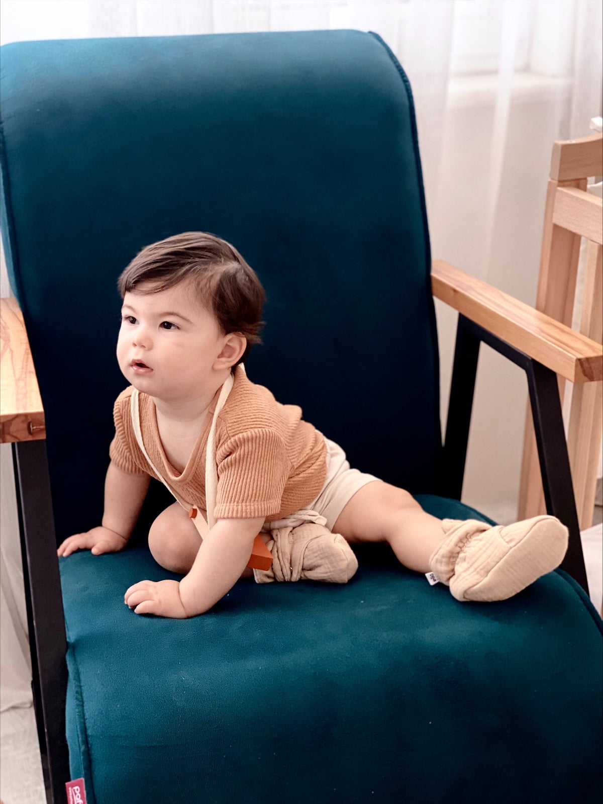 Baby seated sideways in a teal chair wearing beige muslin booties.