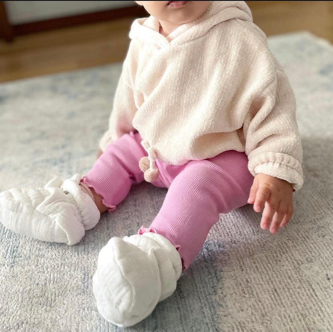 Baby seated on a rug wearing cream booties and pink trousers, front view.