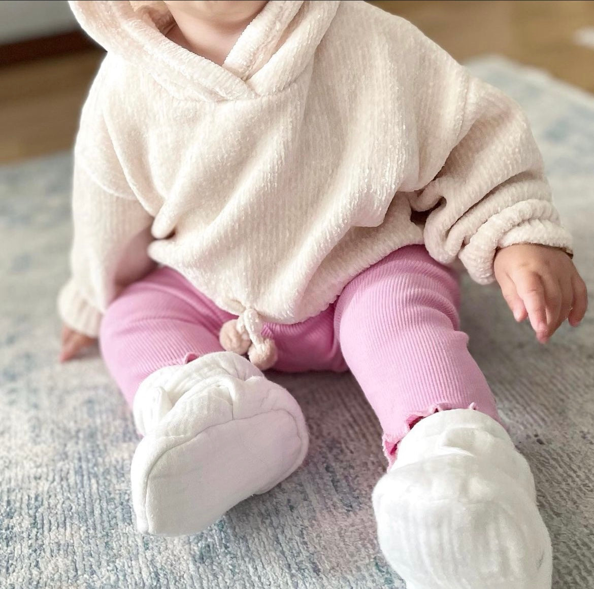 Close-up of cream booties worn by a baby seated on a rug.