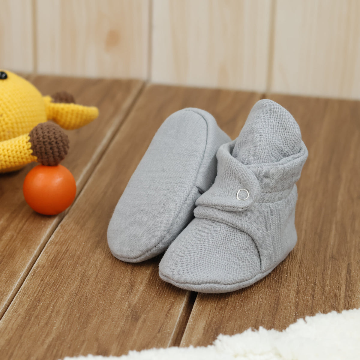 Light gray textured muslin baby booties with snap straps arranged on a surface with one sole facing the camera.