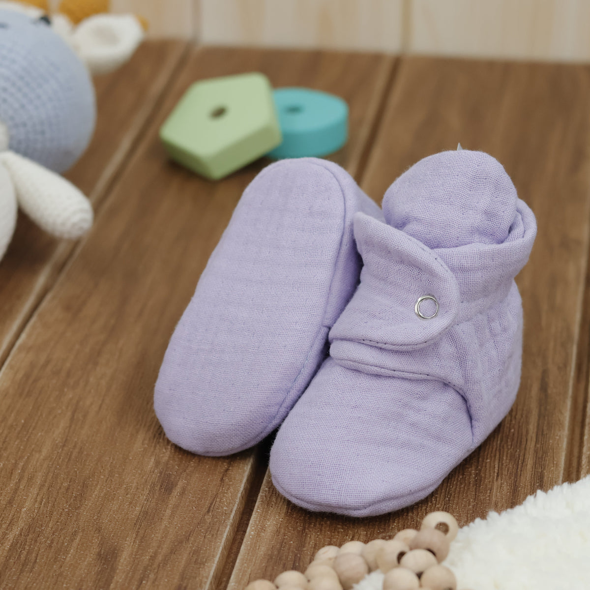 Lilac textured muslin baby booties with snap straps arranged on a surface with one sole facing the camera.