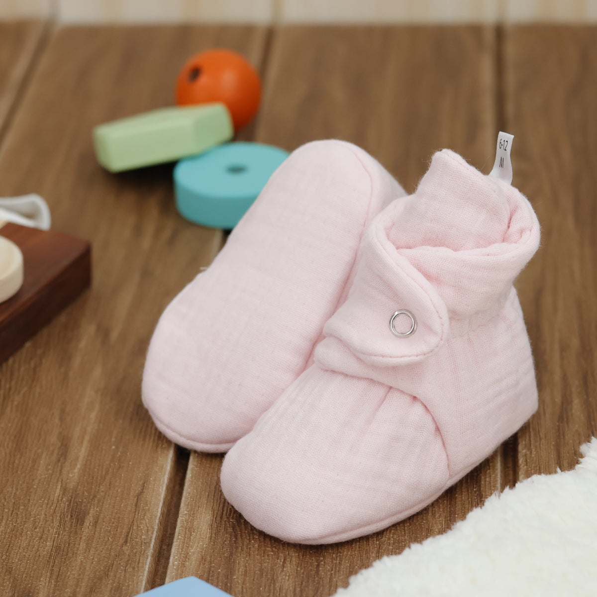 Pale pink textured muslin baby booties with snap straps arranged on a surface with one sole facing the camera.