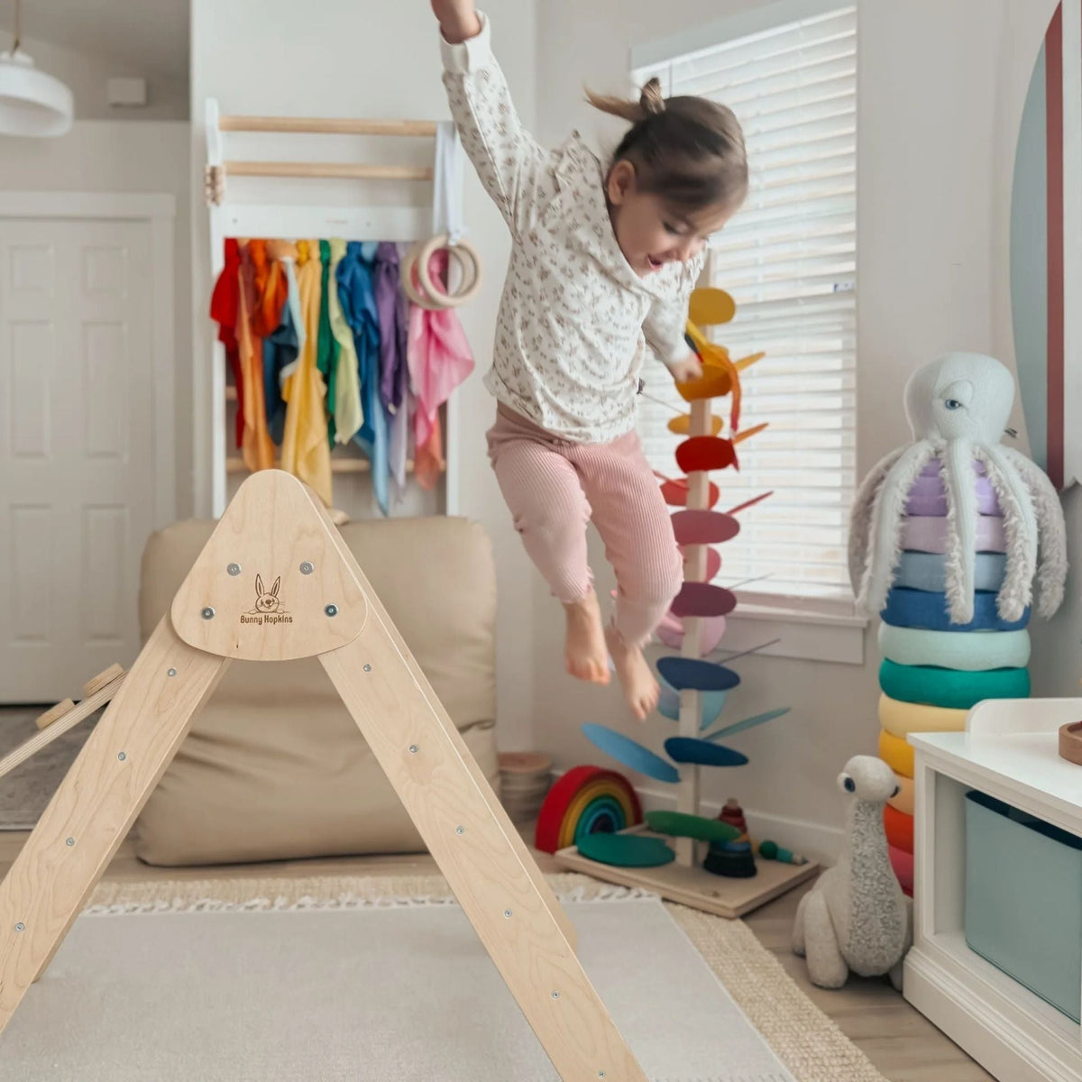 Child airborne beside a pale natural-tone climbing triangle in a playroom.