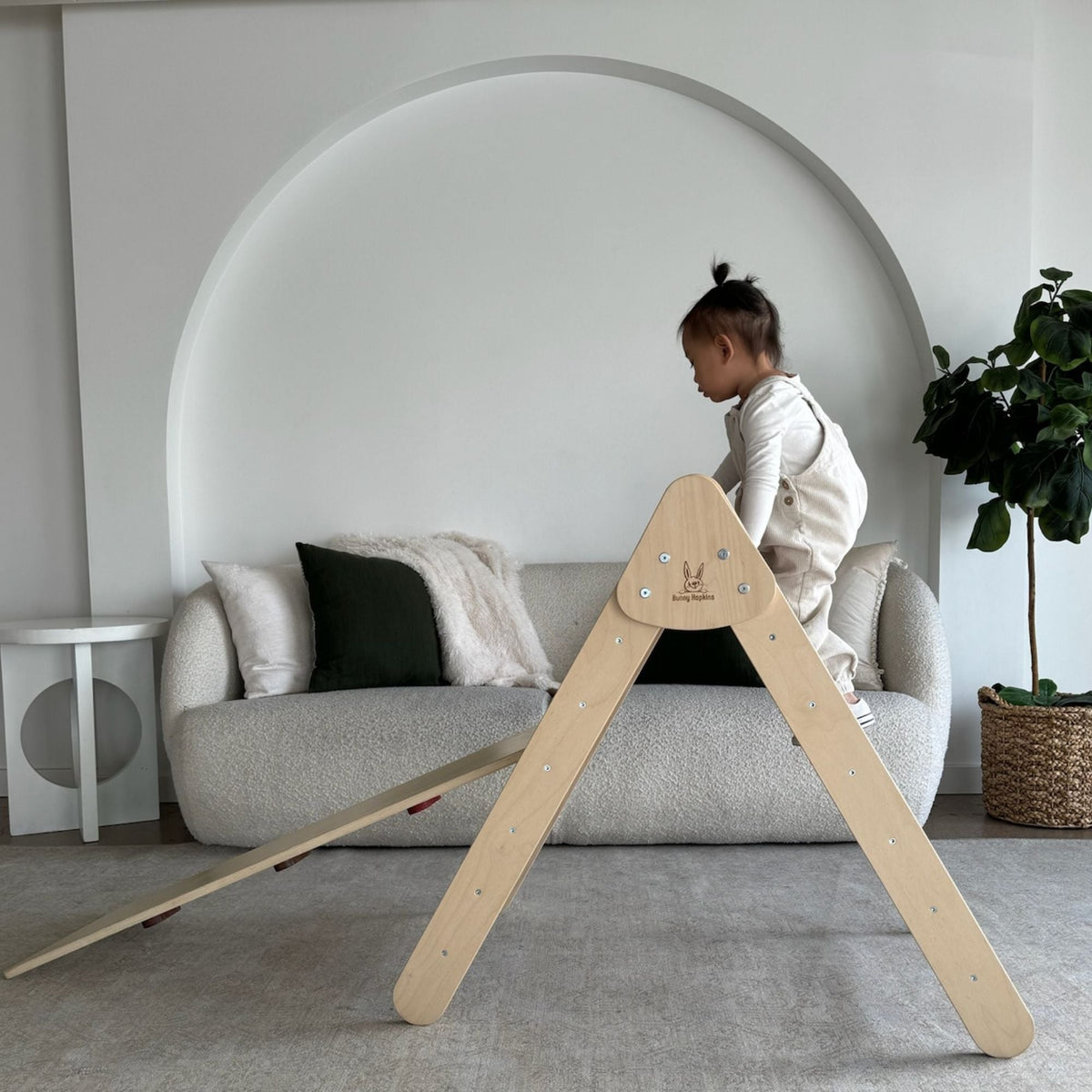 Child standing at the top of a pale climbing triangle with an attached ramp beside a sofa.