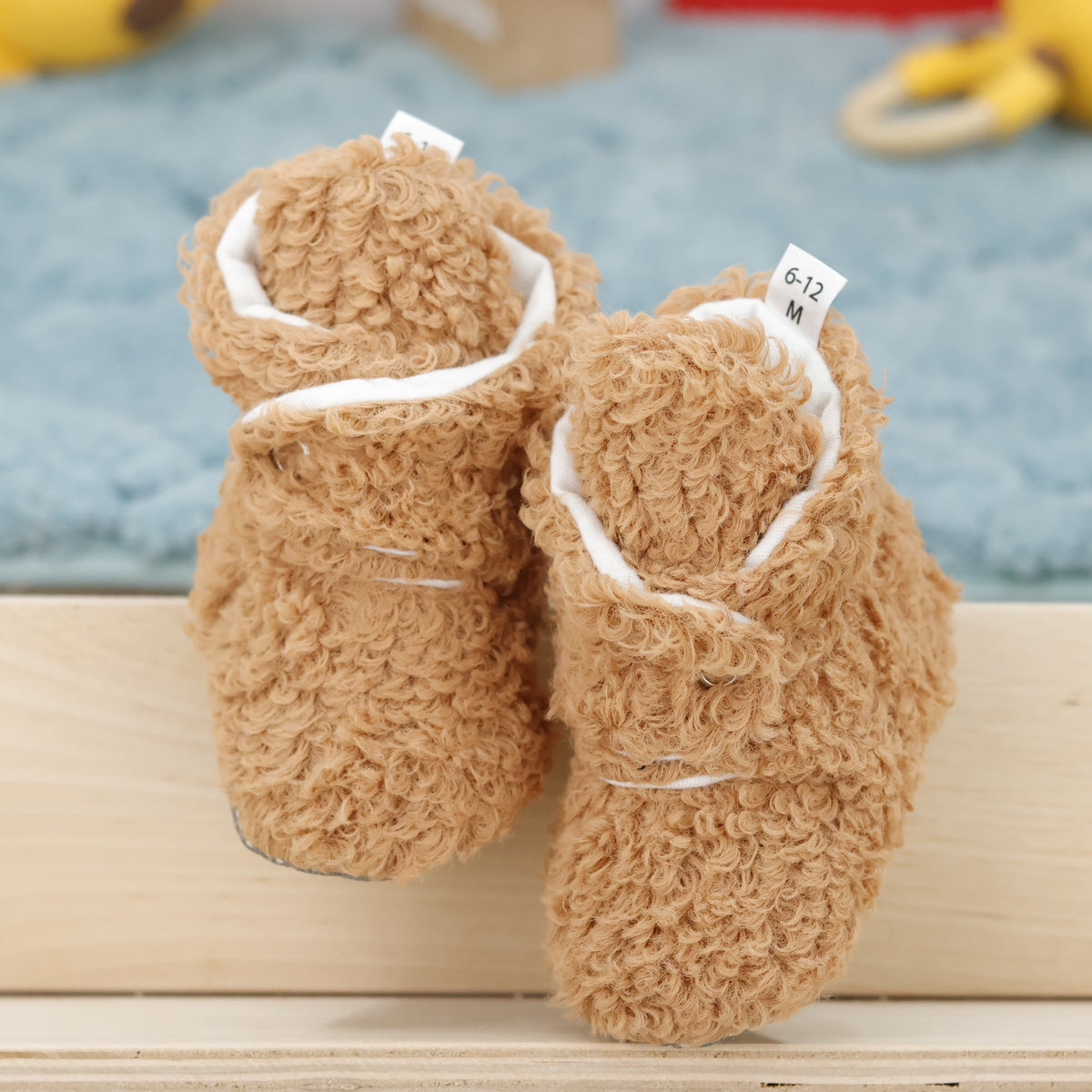 Front view of a pair of light brown teddy-texture baby booties with snap straps and gray grip soles.